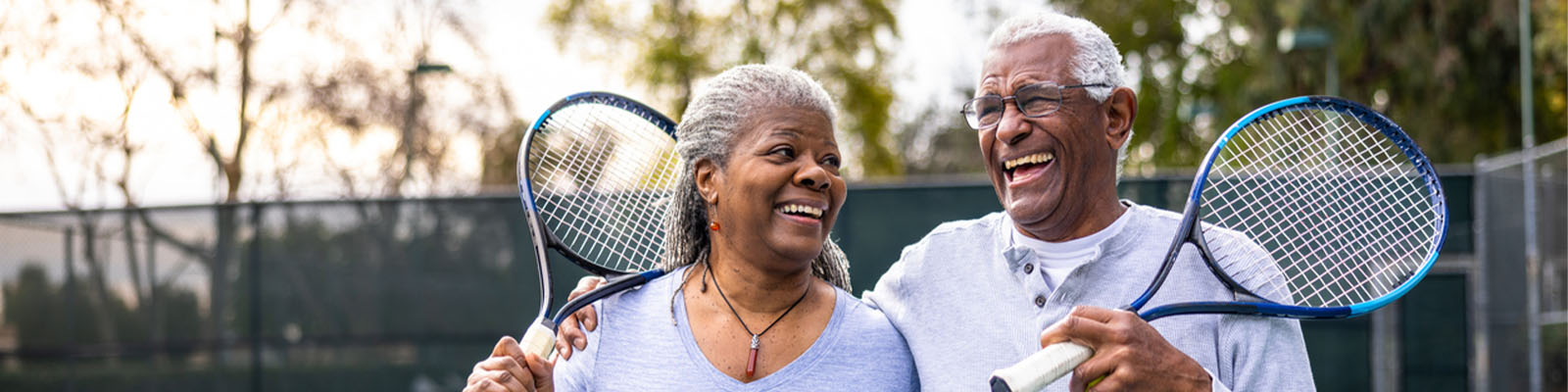 A senior man and woman walking off of a tennis court while holding tennis rackets