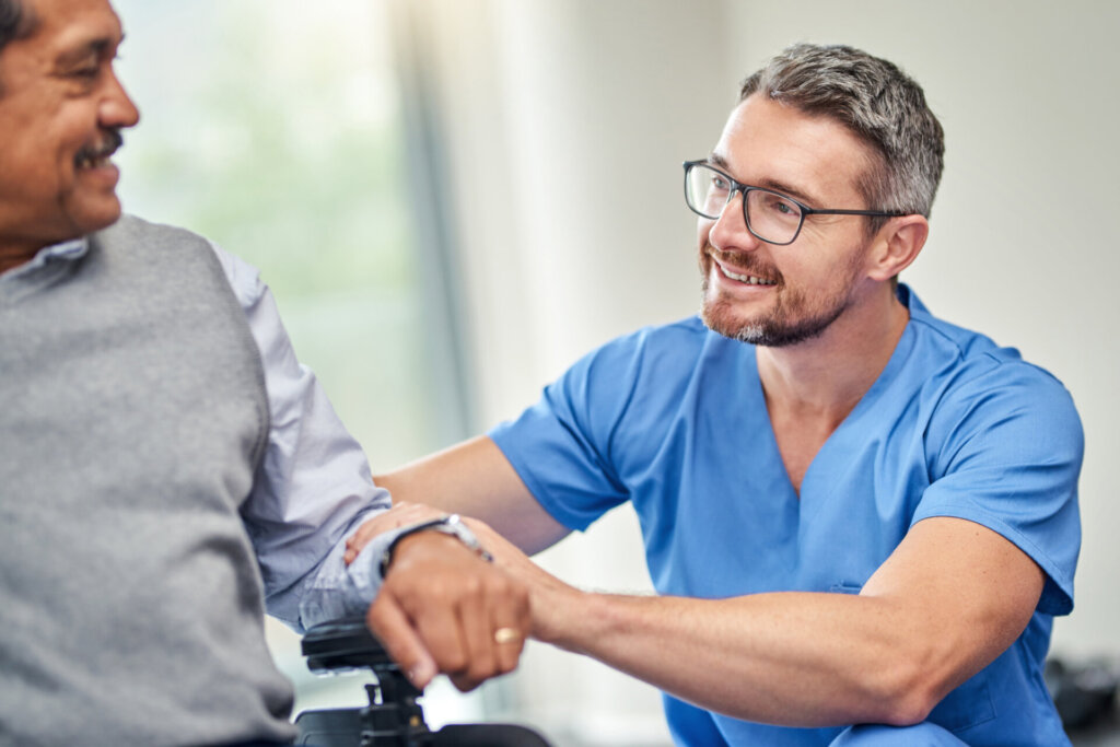 A senior man sitting in a wheelchair as his male assisted living nurse kneels beside him with a smile