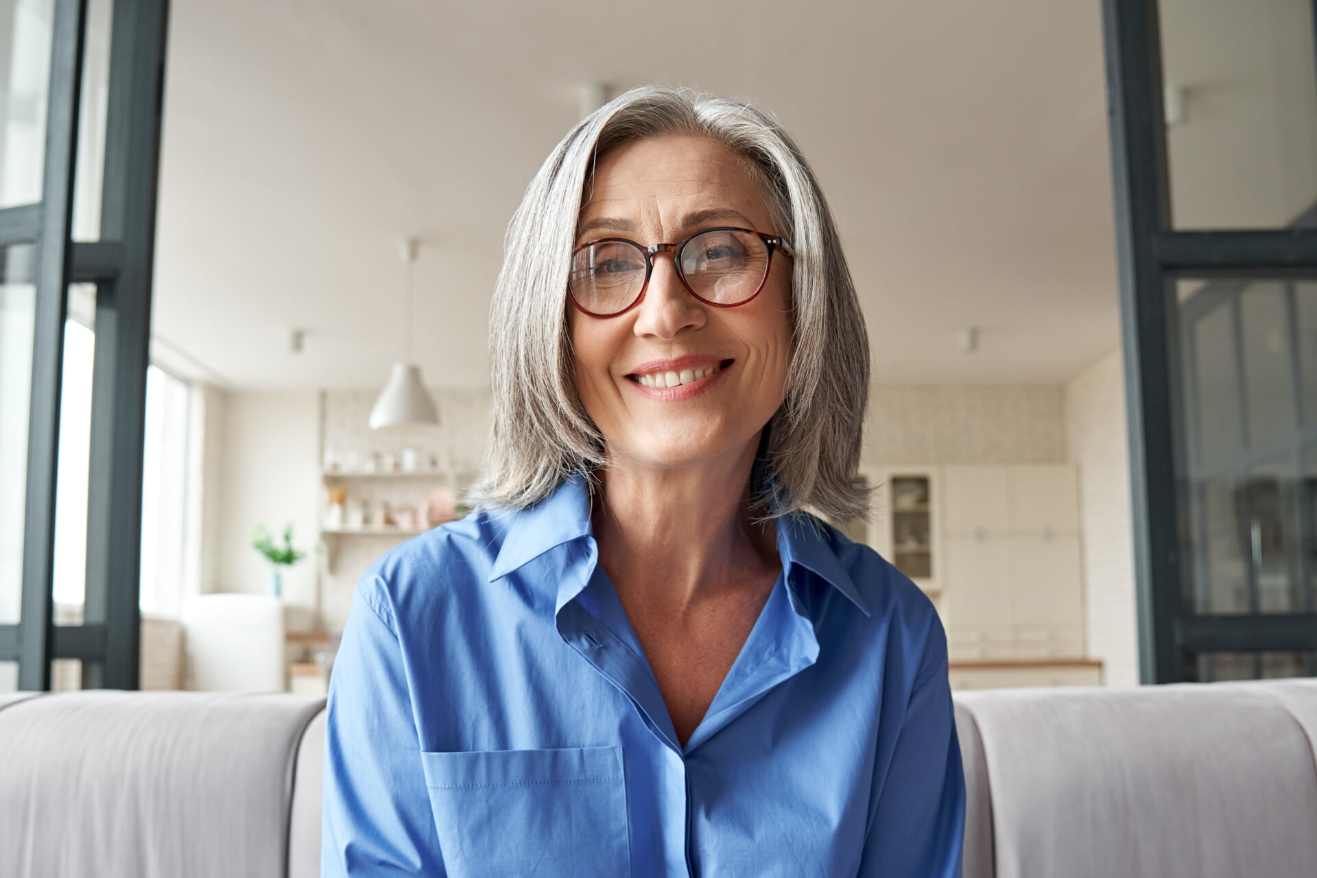 A woman wearing a blue shirt and glasses smiling while sitting on a grey couch